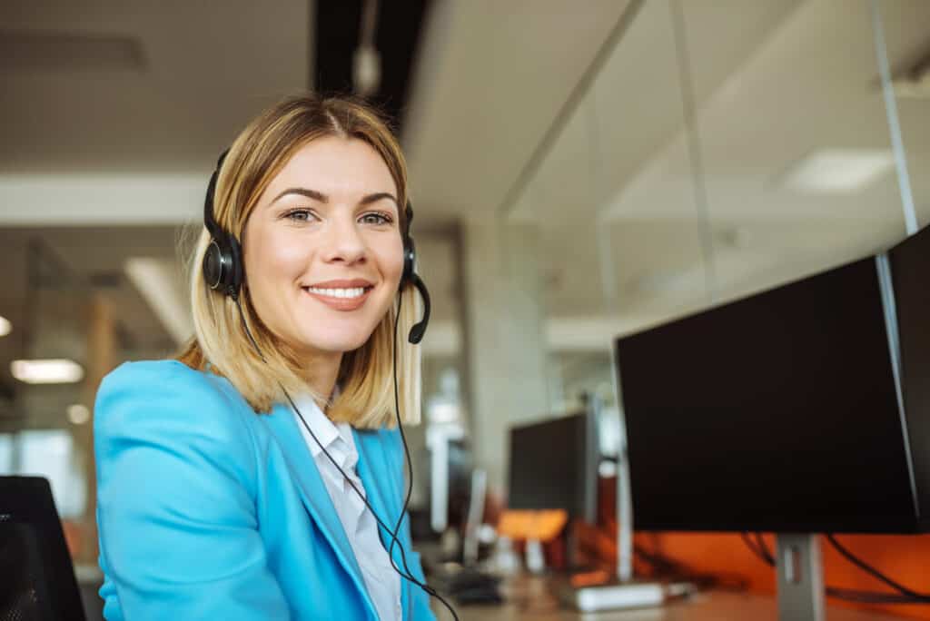 Young female travel agent, wearing a headset and bright blue suit, smiles at the camera from her desk in the office.